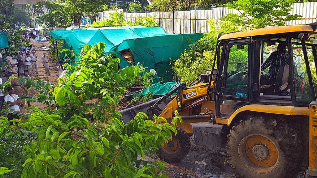 Hyderabad Hydraa demolition drive at Jubilee Hills; a JCB earthmover clearing illegal encroachments covered with green tarpaulins, with police and locals gathered at the site amidst trees and debris.