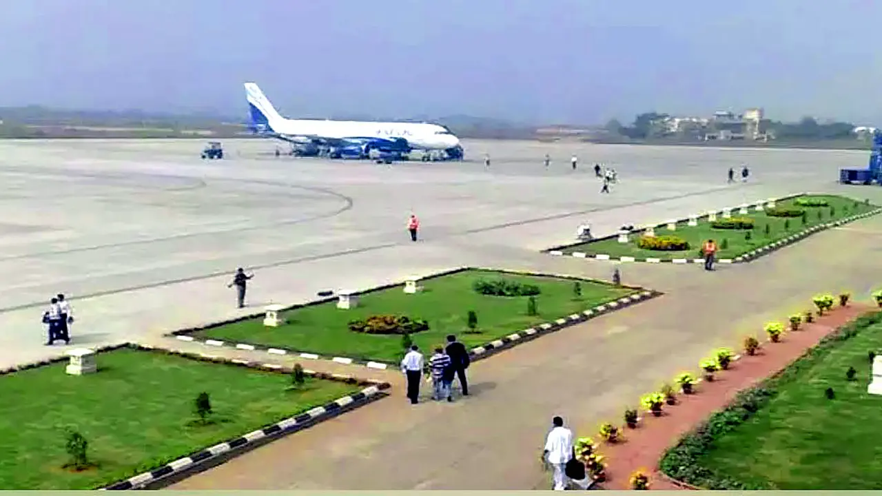 A wide-angle view of Warangal airport tarmac with a parked passenger aircraft, green landscaped areas, ground staff, and visitors walking near the runway.