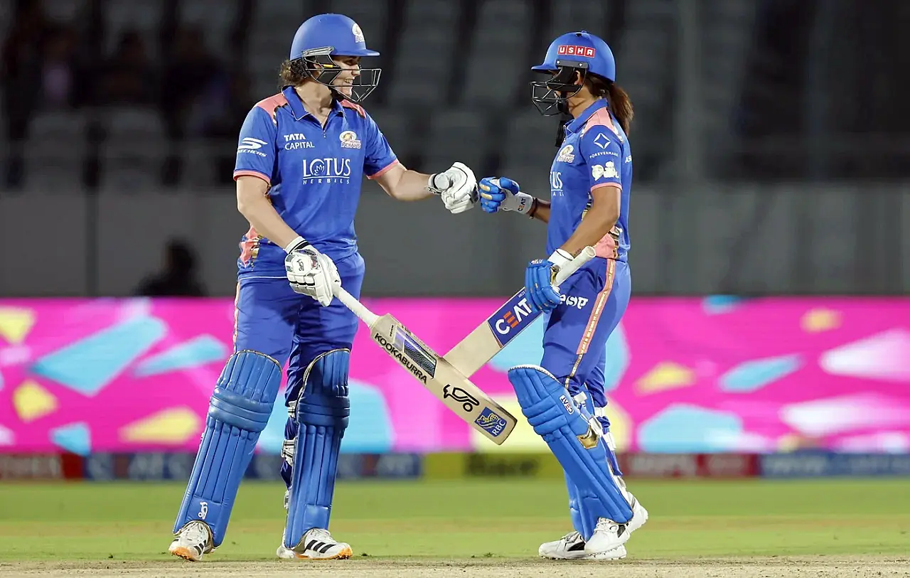 WPL మ్యాచ్‌లో ముంబై ఇండియన్స్ ఆటగాళ్లు నాట్ స్కైవర్–బ్రంట్, హర్మన్‌ప్రీత్ కౌర్ మధ్యలో రన్ తీసుకున్న తర్వాత ఒకరికొకరు గ్లౌవ్స్ తాకిస్తూ సెలబ్రేట్ చేస్తున్న దృశ్యం.