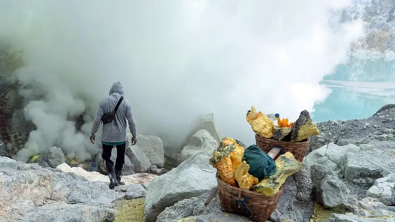 Sulfur baskets filled with yellow sulfur chunks near a miner walking through thick volcanic smoke at Kawah Ijen