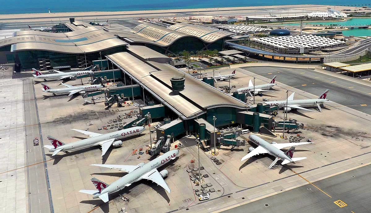 Aerial view of Hamad International Airport in Doha, Qatar, showing multiple Qatar Airways wide-body aircraft (including Boeing 777 and Airbus A350 models in white with maroon/red tail) parked at gates along the terminal building, with the turquoise sea and airport infrastructure in the background. Flights grounded due to Qatar's airspace closure.